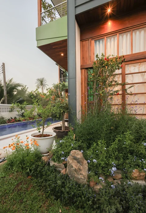 Villa garden at dusk with flower beds and warm lights glowing on the wooden facade, pool beyond