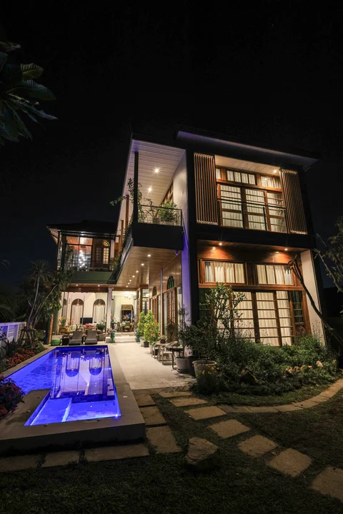 Dramatic night shot of the villa with blue pool glowing, two-storey lit facade and tropical garden