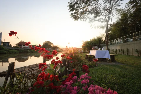 Garden riverbank with red and pink flowers, bamboo raft dock and dining table at sunset