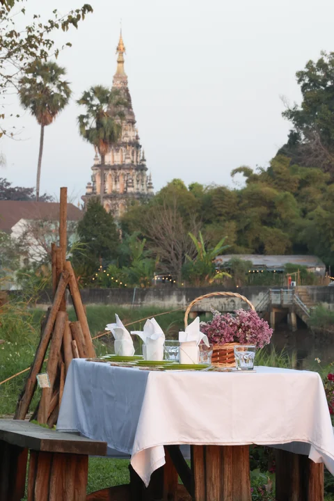 Riverside dining table set for two with flower basket, ancient Buddhist chedi visible across the river